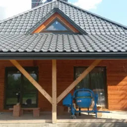 Dom z drewnianą elewacją i grafitowym dachem z dachówki, widoczna betoniarka i elementy konstrukcyjne werandy w trakcie budowy.