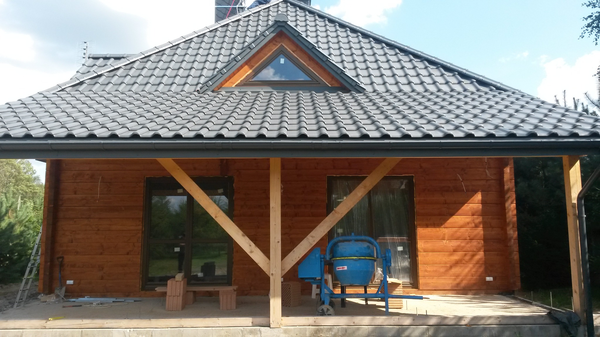 Dom z drewnianą elewacją i grafitowym dachem z dachówki, widoczna betoniarka i elementy konstrukcyjne werandy w trakcie budowy.