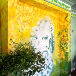 aleksander skrok art supplier - Żółto-zielona ściana z graffiti przedstawiającym twarz brodatego mężczyzny w stylu greckim, z napisem 'ODINO DAL 1965', dodatkowo ozdobiona motywem cytryn na górze i drzewkiem cytrynowym w doniczce...