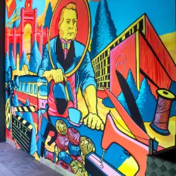 Barwny mural w stylu pop-art przedstawiający postać w owalnej ramie, tramwaj, budynek z napisem FUZJA, nici, nożyczki, maszynę do szycia i inne elementy związane z przemysłem włókienniczym.