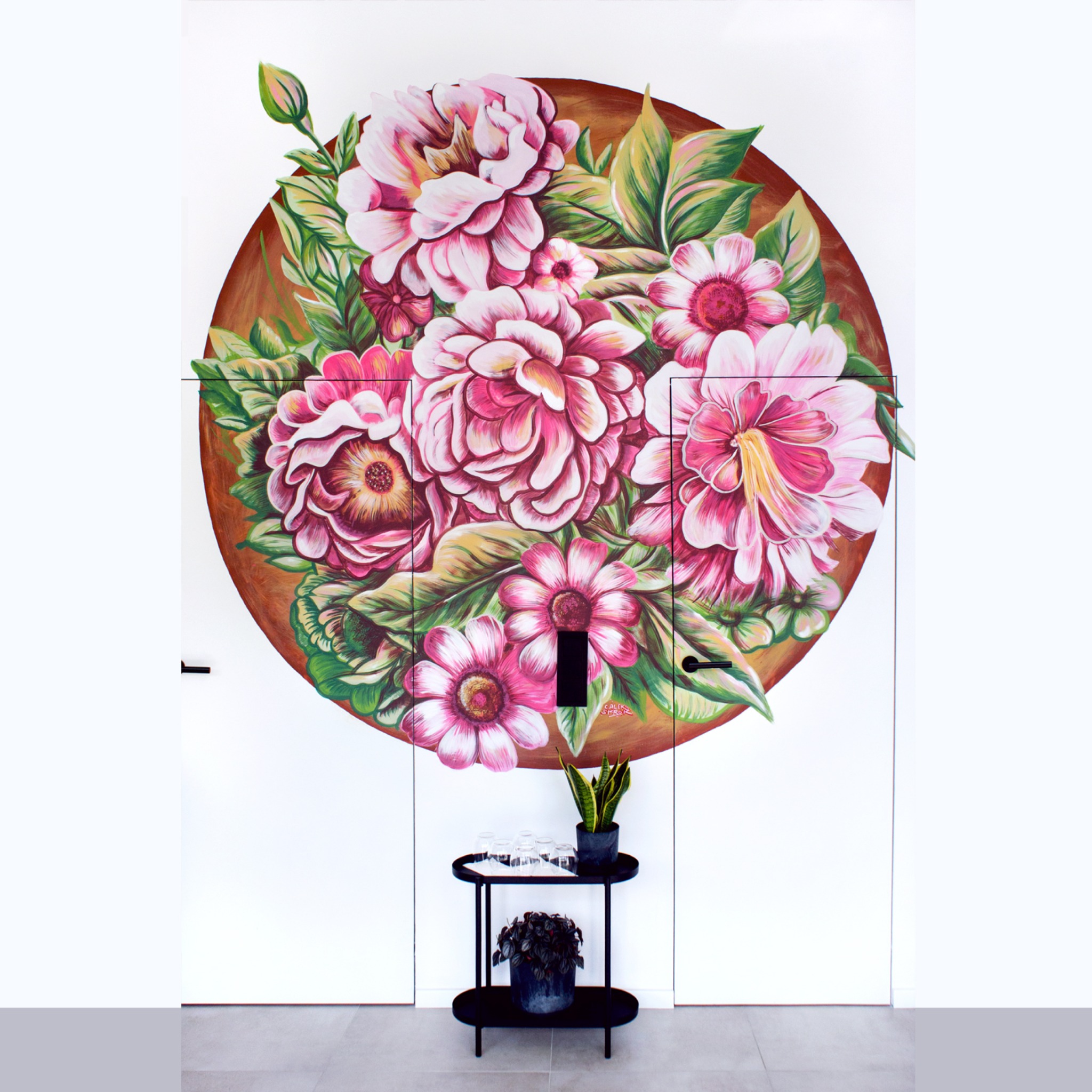 Ręcznie malowana kompozycja kwiatowa w okręgu na białych drzwiach szafy wnękowej, z roślinami doniczkowymi i szklankami na czarnym stoliku na pierwszym planie.