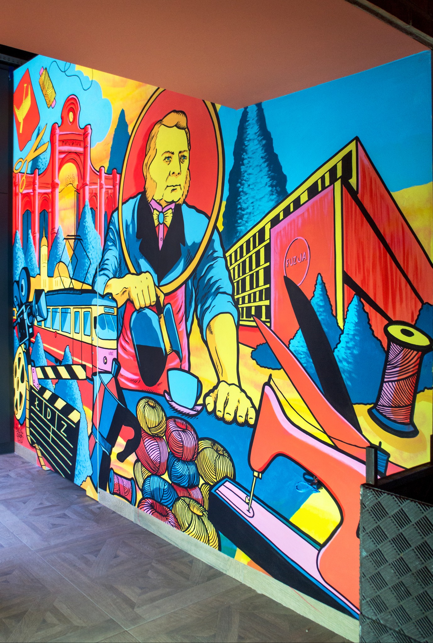 Barwny mural w stylu pop-art przedstawiający postać w owalnej ramie, tramwaj, budynek z napisem FUZJA, nici, nożyczki, maszynę do szycia i inne elementy związane z przemysłem włókienniczym.