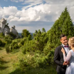 Para młoda w objęciach na tle malowniczego krajobrazu ze skałami i zielenią, on w czarnym garniturze z bordową muchą, ona w białej sukni z koronkowymi rękawami.