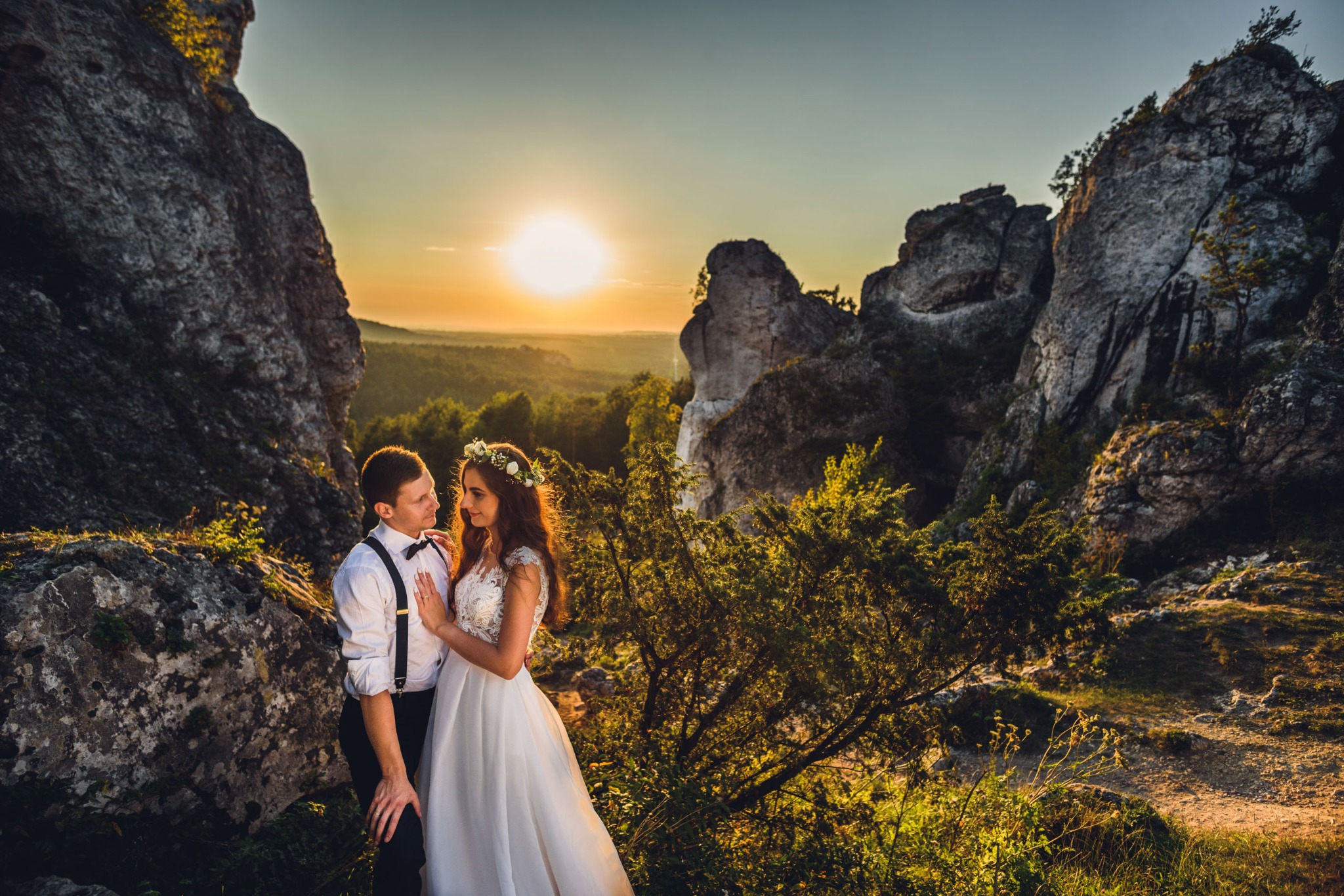 Para młoda w objęciach na tle skalistego krajobrazu i zachodzącego słońca, panna młoda w białej sukni z koronkową górą i wiankiem, pan młody w białej koszuli z muchą i szelkami.