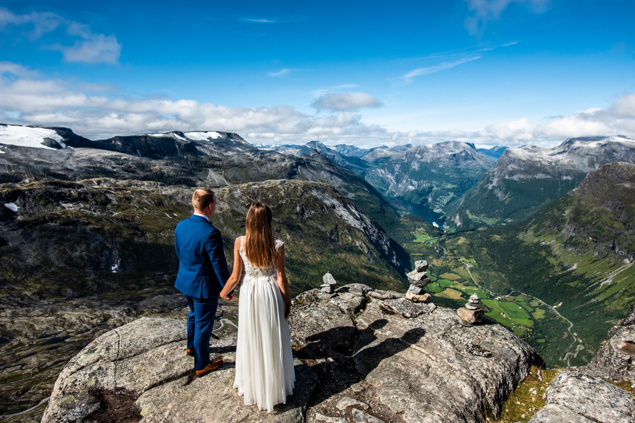Para młoda trzymająca się za ręce na skalistym szczycie góry z widokiem na dolinę i góry w tle, dzień ślubu w plenerze.