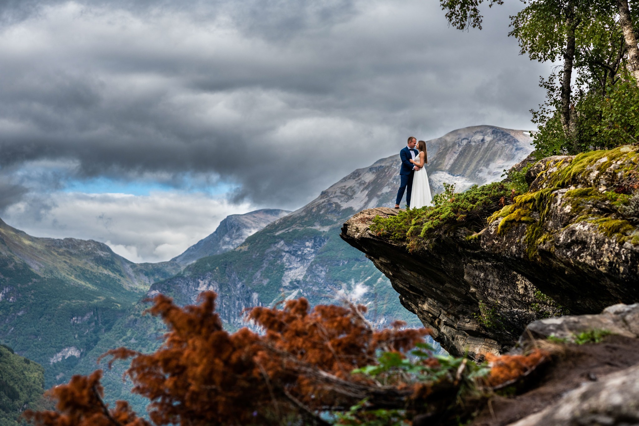 Para młoda w objęciach na skalnym klifie z widokiem na góry i zachmurzone niebo, on w granatowym garniturze, ona w białej sukni ślubnej.