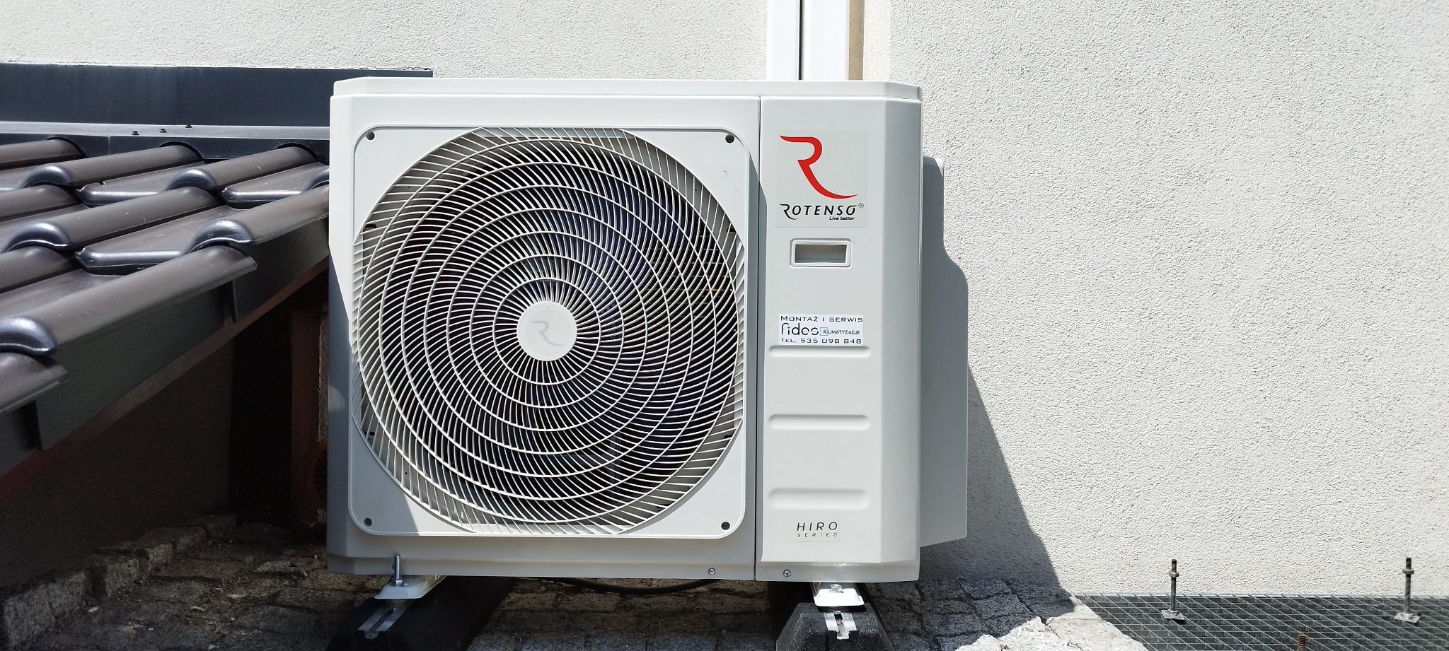 Zewnętrzna jednostka klimatyzacji Rotenso Hiro Series zamontowana na wspornikach przy ścianie budynku z widoczną strukturą tynku i częściowo pod okapem dachu pokrytego dachówką.