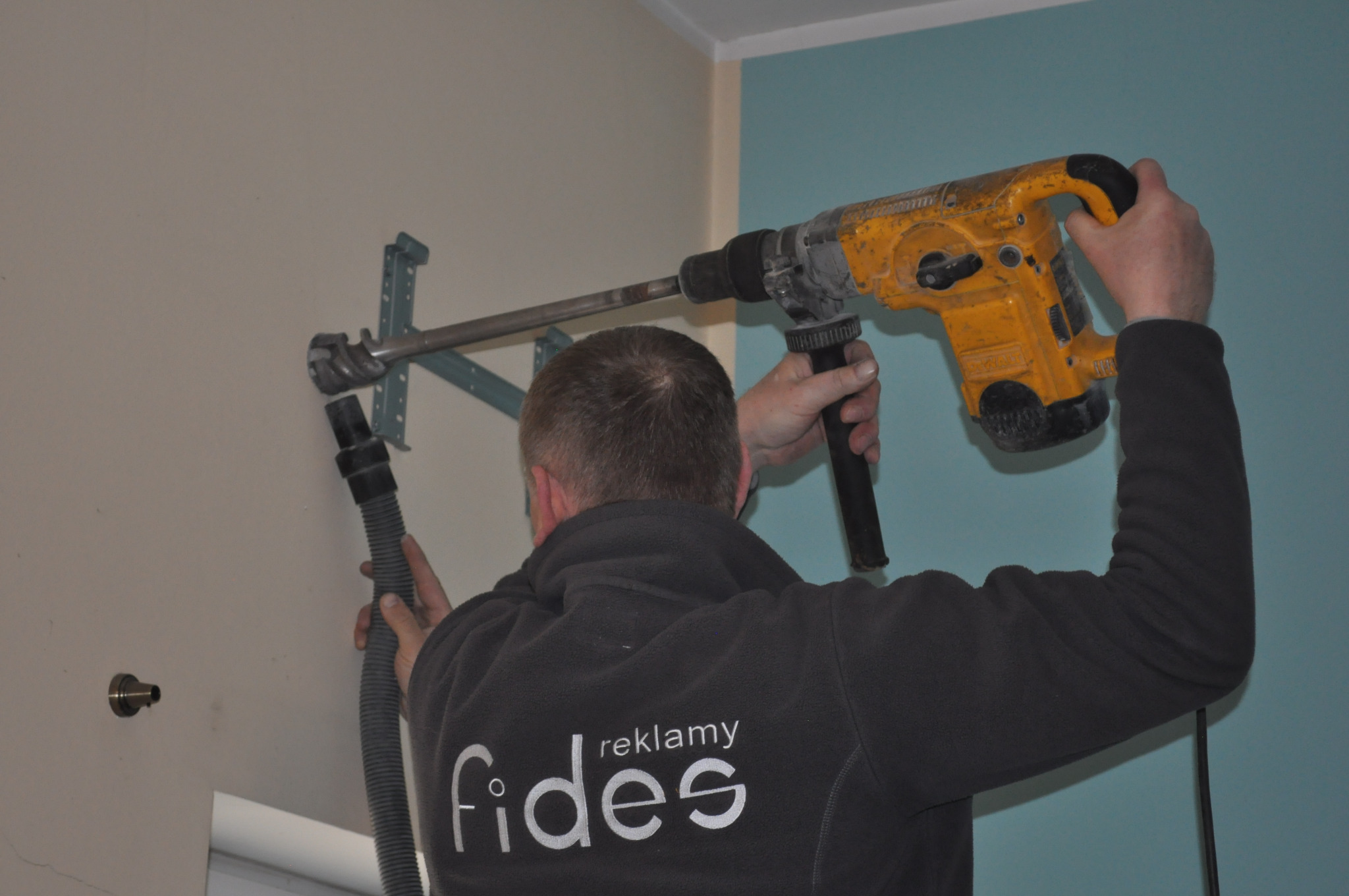 Instalator wierci otwór w ścianie pod montaż klimatyzacji za pomocą wiertarki udarowej z odciągiem pyłu, widoczne elementy mocujące. Na bluzie logo firmy Fides Reklamy.