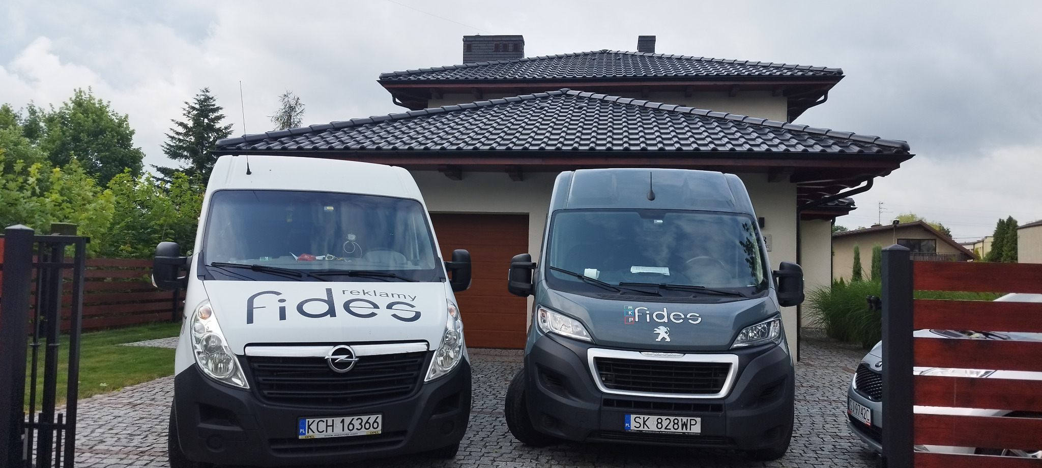 Dwa samochody dostawcze firmy Fides Reklamy zaparkowane przed nowoczesnym domem z ciemną dachówką, widoczne tablice rejestracyjne i logo firmy na pojazdach.