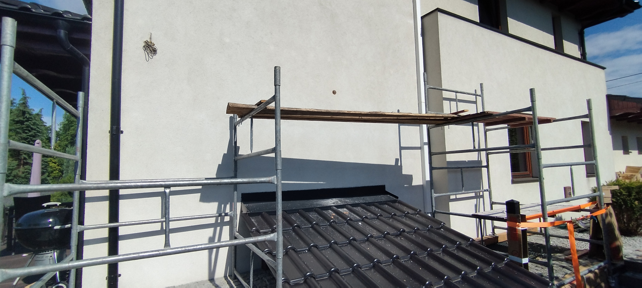 Dom jednorodzinny w trakcie ocieplania, widoczne rusztowanie z drewnianymi pomostami, czarny dach z dachówki ceramicznej, biała elewacja budynku.