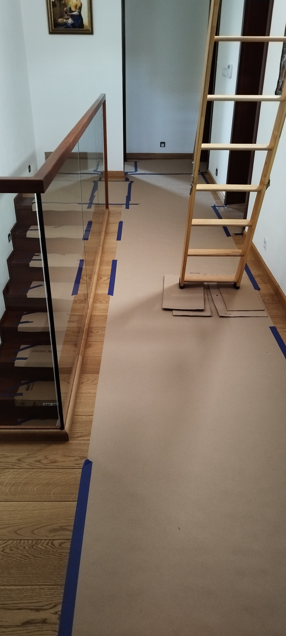 Przygotowanie do montażu klimatyzacji w domu: podłoga i schody zabezpieczone kartonem i niebieską taśmą malarską, drabina oparta o ścianę.