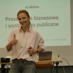 Uśmiechnięta trenerka prezentuje kciuk w górę na tle ekranu z napisem 'Prezentacje biznesowe i wystąpienia publiczne', na stole laptop i środek dezynfekujący.