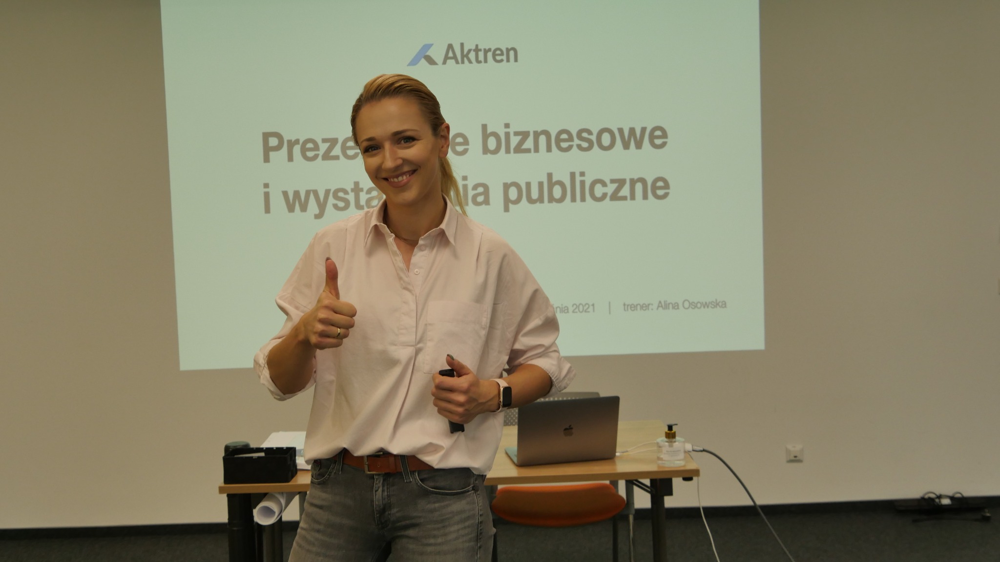 Uśmiechnięta trenerka prezentuje kciuk w górę na tle ekranu z napisem 'Prezentacje biznesowe i wystąpienia publiczne', na stole laptop i środek dezynfekujący.
