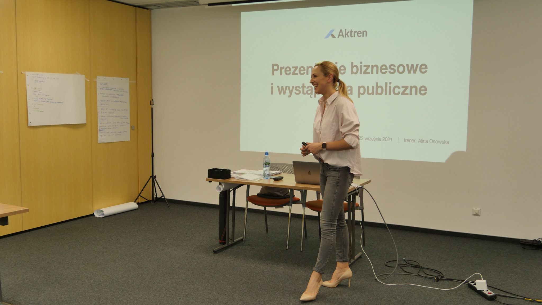 Kobieta w jasnej koszuli i szarych jeansach, uśmiechnięta, prezentuje szkolenie z prezentacji biznesowych i wystąpień publicznych, widoczny ekran z logo firmy Aktren i datą 22 września 2021...