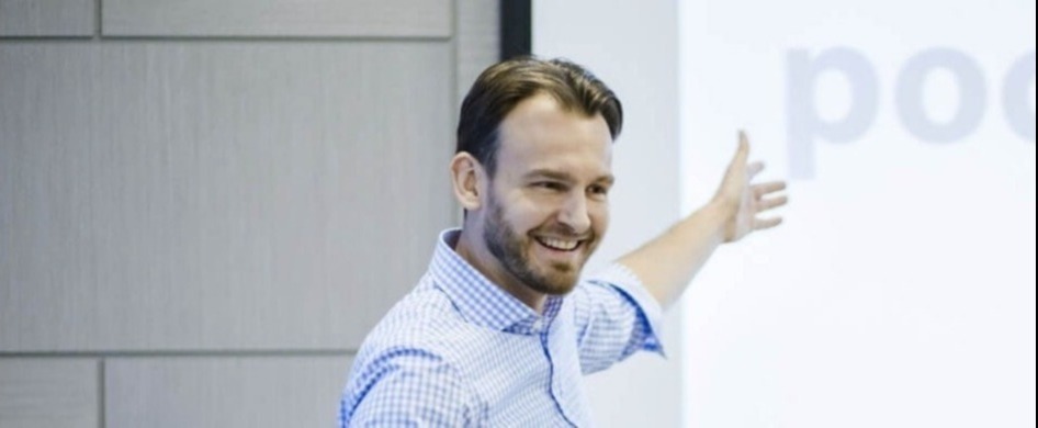 Uśmiechnięty mężczyzna w niebieskiej koszuli w kratę prezentuje slajd z napisem podczas kursu zawodowego.