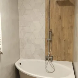 Nowoczesna łazienka z wolnostojącą wanną na nóżkach, drewnopodobnymi płytkami ściennymi i podłogowymi oraz geometrycznymi płytkami nad wanną.