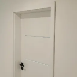 Zamknięte, białe drzwi wewnętrzne z poziomymi, wąskimi wstawkami i czarną klamką na tle białej ściany.