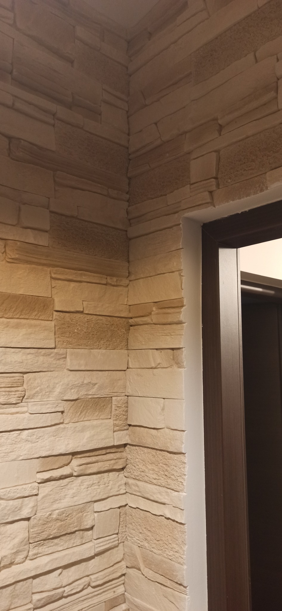 Wewnętrzny narożnik pomieszczenia wykończony dekoracyjną okładziną ścienną imitującą kamień w odcieniach beżu, sąsiadujący z ościeżnicą drzwiową w kolorze ciemnego brązu.