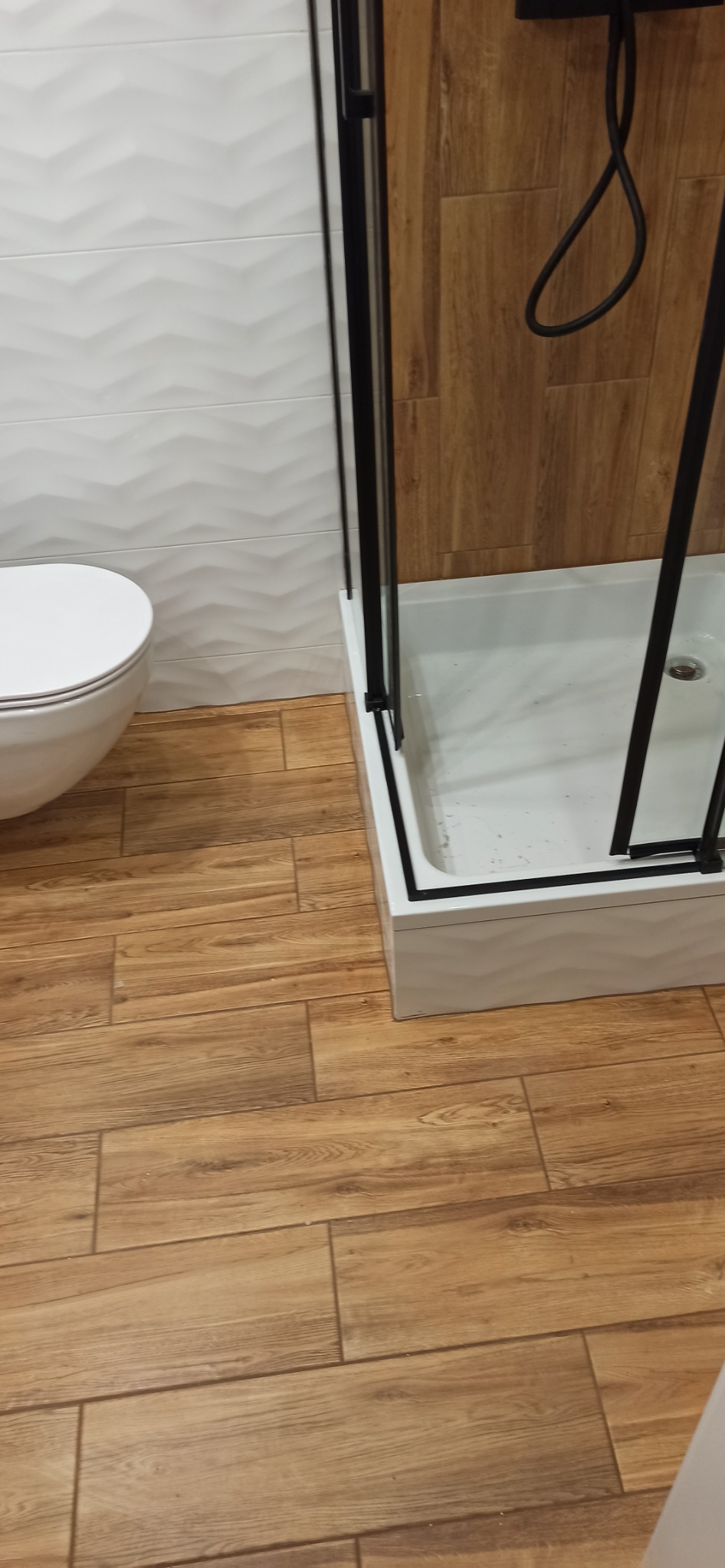 Nowoczesna łazienka z kabiną prysznicową z czarnymi ramami, białą miską WC i podłogą imitującą drewno, uzupełnona białą ścianą z delikatnym wzorem 3D.