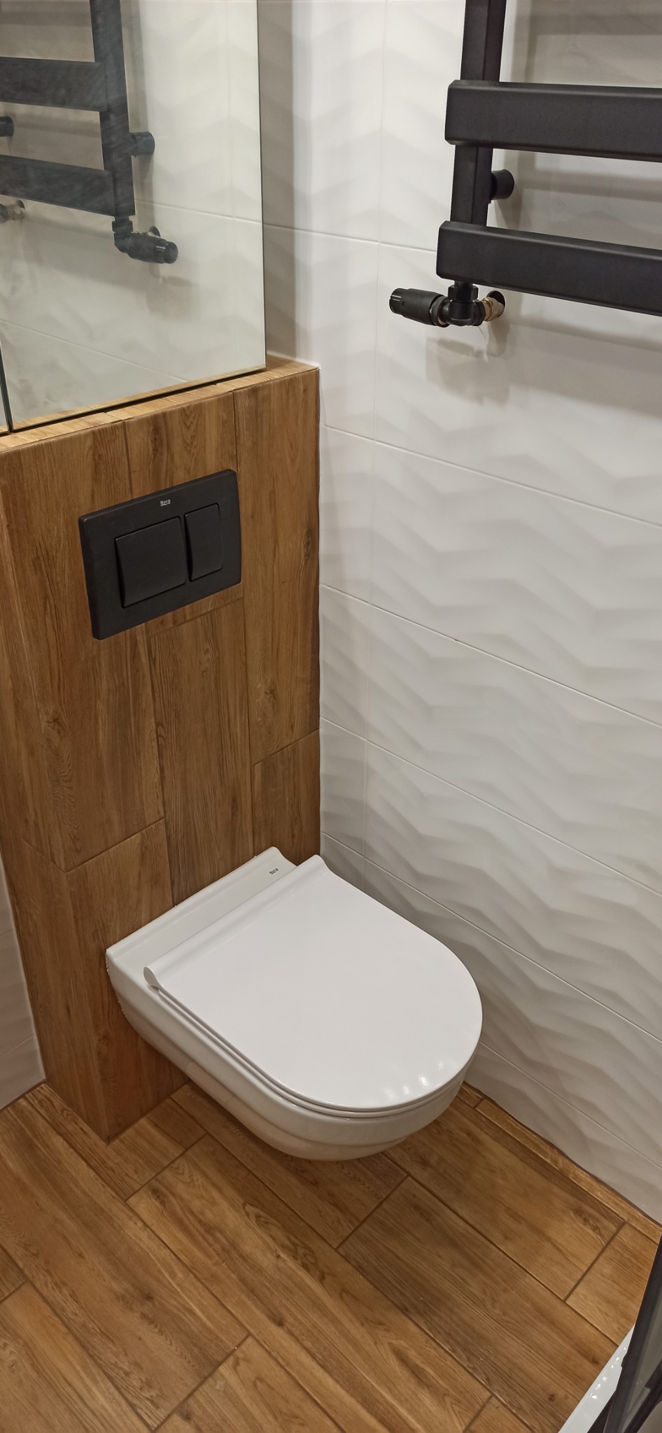 Nowoczesna toaleta podwieszana z czarnym przyciskiem spłukującym na drewnianej obudowie, białe płytki ścienne z delikatnym wzorem, drewnopodobna podłoga i czarny grzejnik drabinkowy.