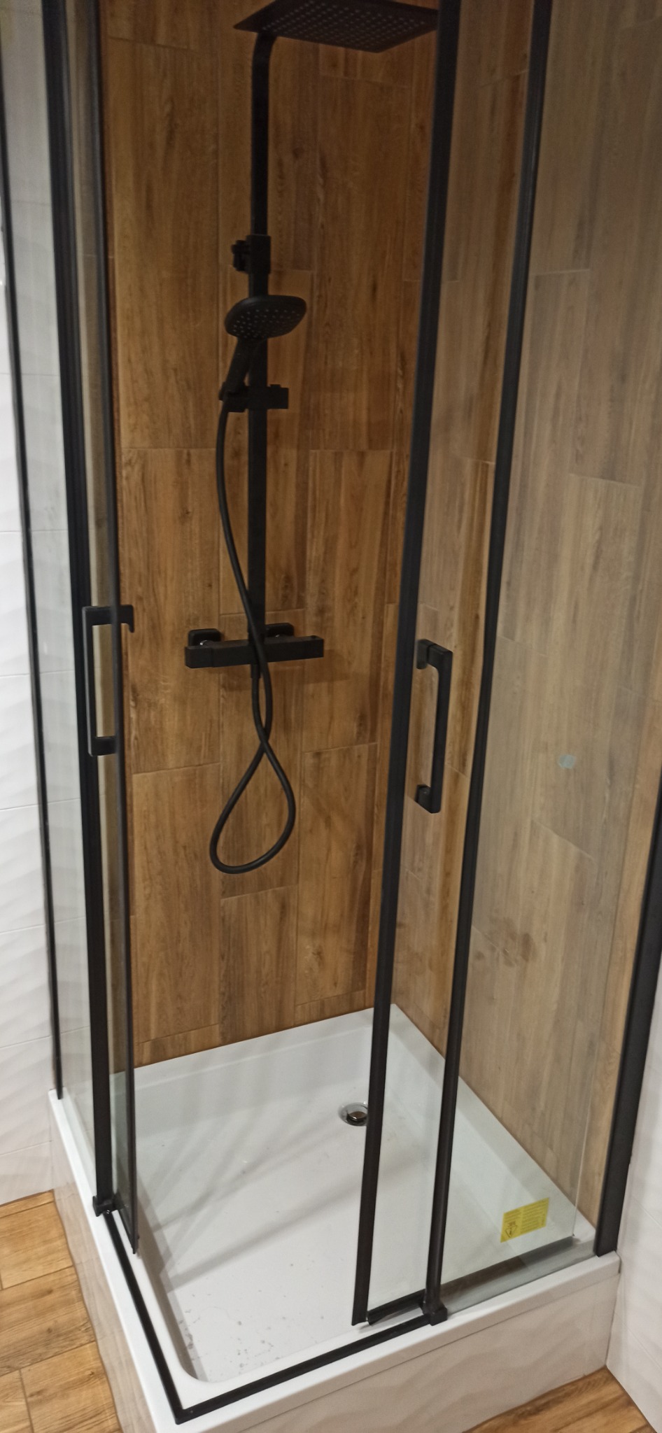 Nowoczesna kabina prysznicowa z czarnymi ramami i deszczownicą, wykończona płytkami imitującymi drewno i białymi płytkami.