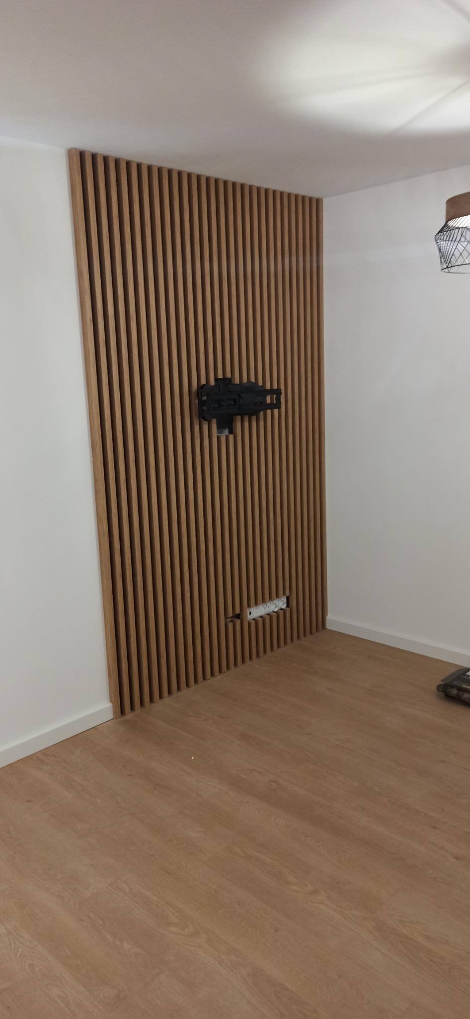 Panel ścienny z drewnianych listewek z zamontowanym uchwytem do telewizora i gniazdkami elektrycznymi, jasna podłoga.