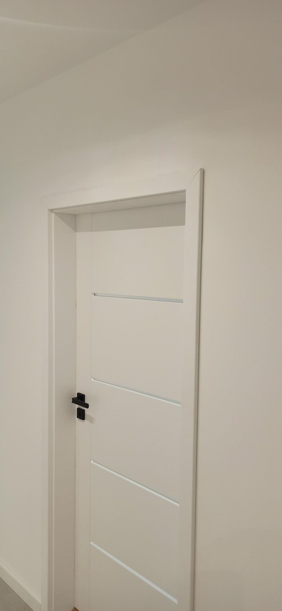 Zamknięte, białe drzwi wewnętrzne z poziomymi, wąskimi wstawkami i czarną klamką na tle białej ściany.