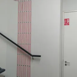 Czerwone przewody alarmowe, równolegle ułożone i przymocowane do białej ściany korytarza obok schodów i drzwi ewakuacyjnych, z widocznymi puszkami połączeniowymi na górze.