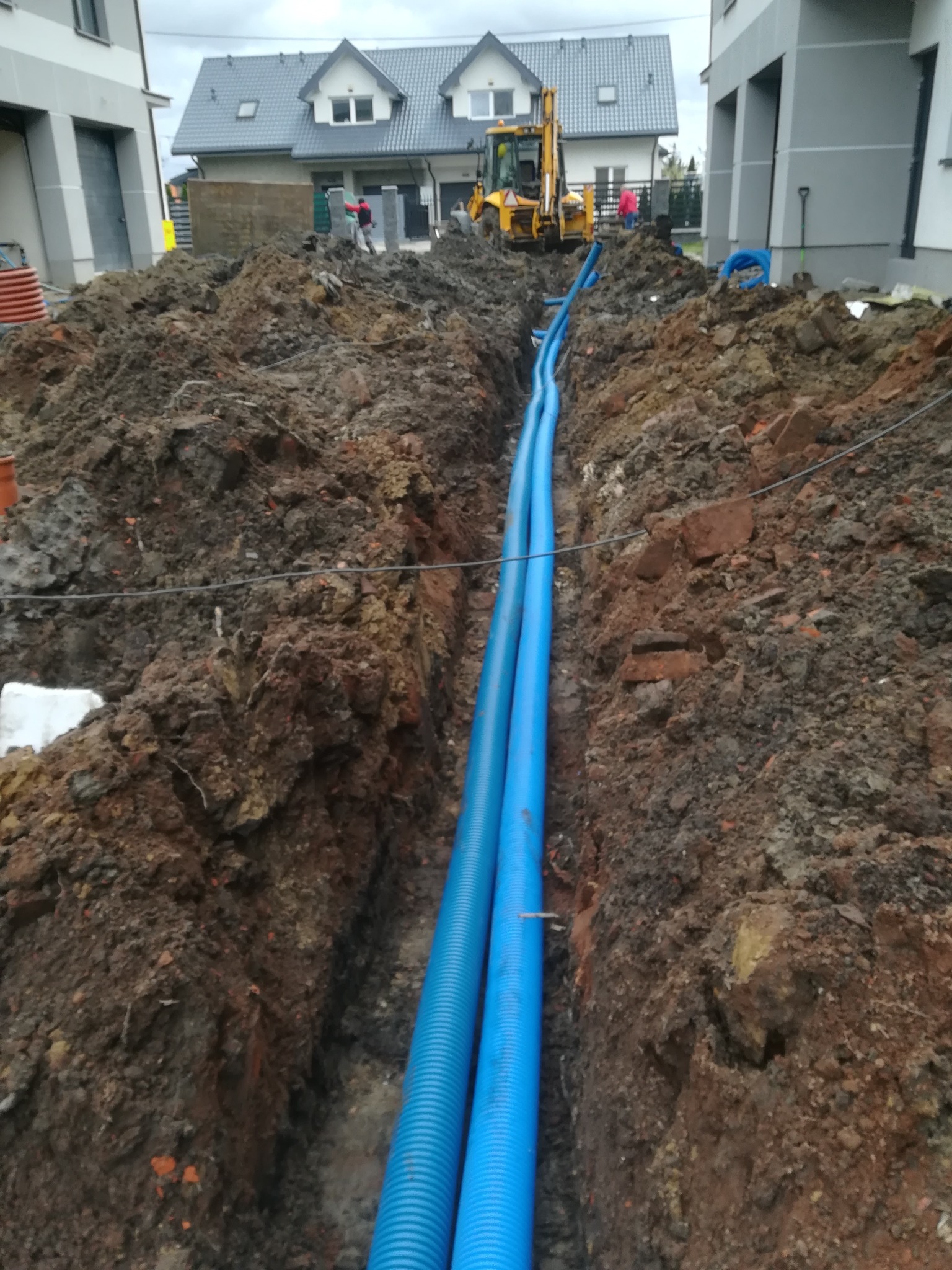Wykop z dwoma niebieskimi, karbowanymi rurami ułożonymi wzdłuż wykopu. W tle widoczny budynek w trakcie budowy oraz koparka.