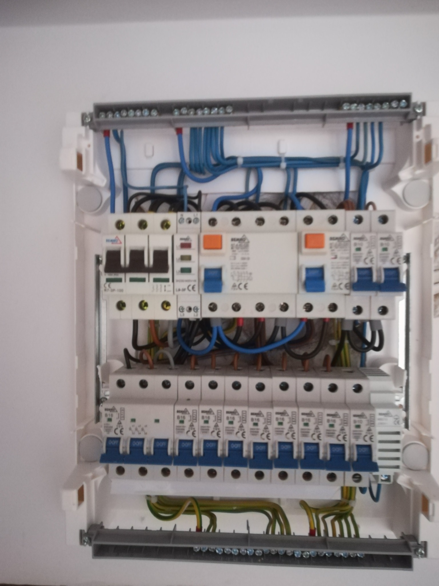 Otwarta skrzynka rozdzielcza z widocznymi bezpiecznikami, przewodami w kolorach niebieskim, czarnym i żółto-zielonym, zamontowana na białej ścianie.