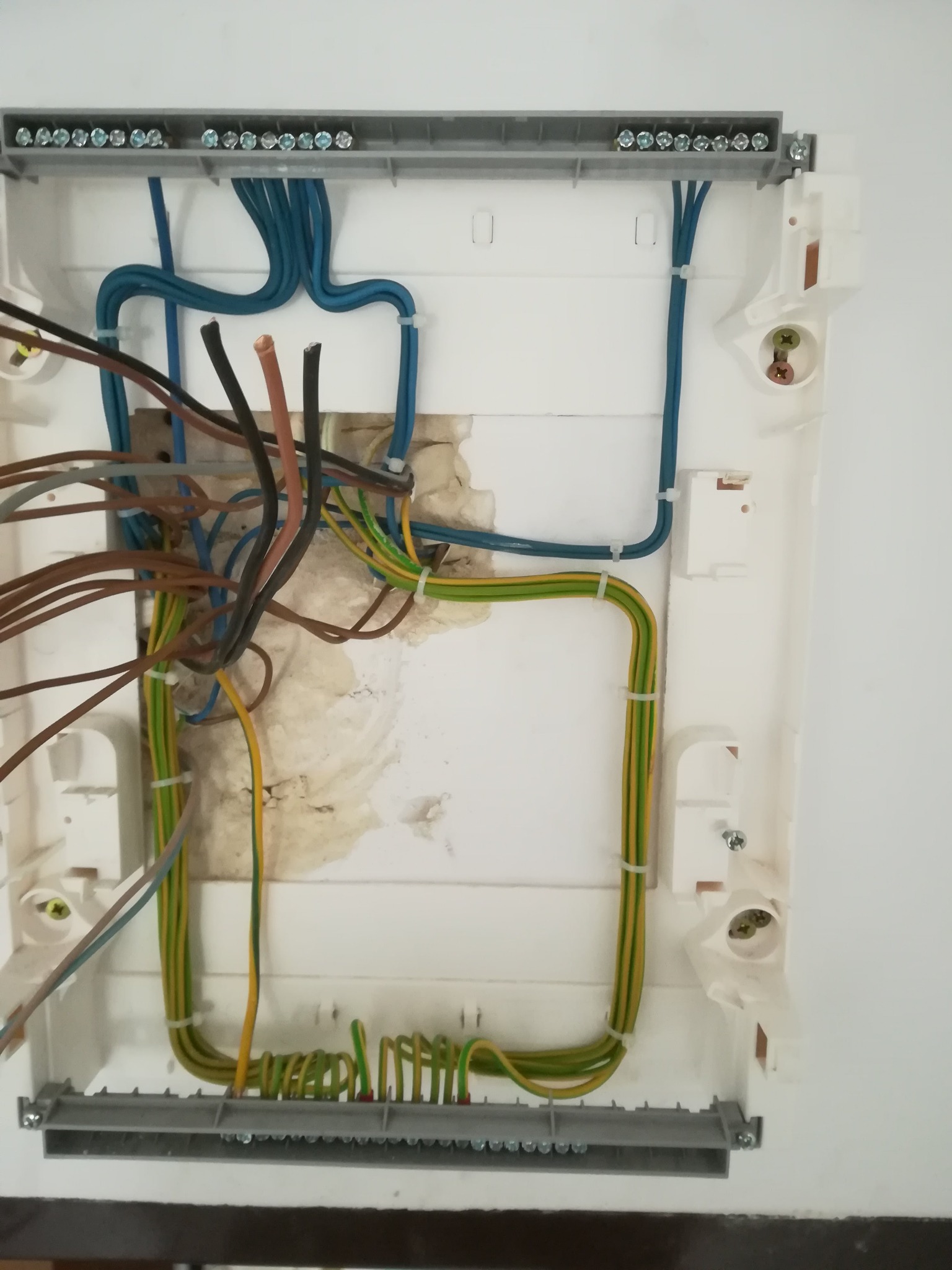 Otwarta biała skrzynka elektryczna z widocznymi przewodami w różnych kolorach (niebieski, brązowy, zielono-żółty) podłączonymi do listew zaciskowych; widoczne ślady po poprzednich instalacjach...