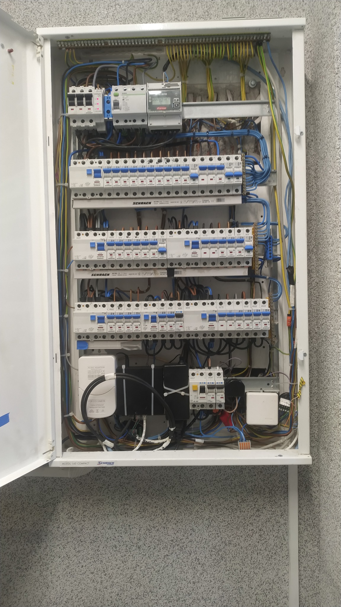 Otwarta skrzynka rozdzielcza z licznikiem energii, bezpiecznikami i okablowaniem wewnątrz, zamontowana na szarej ścianie.