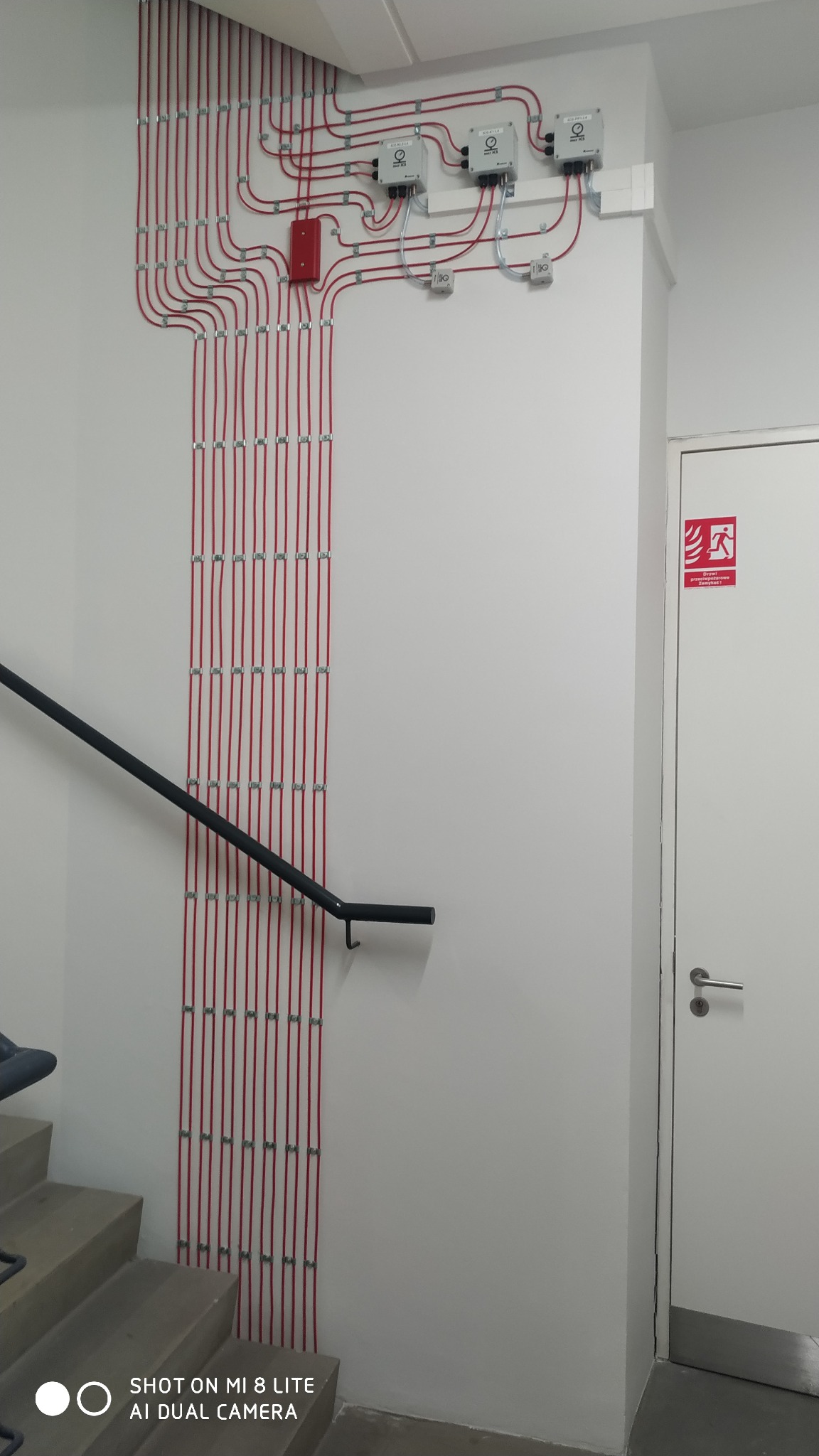 Czerwone przewody alarmowe, równolegle ułożone i przymocowane do białej ściany korytarza obok schodów i drzwi ewakuacyjnych, z widocznymi puszkami połączeniowymi na górze.