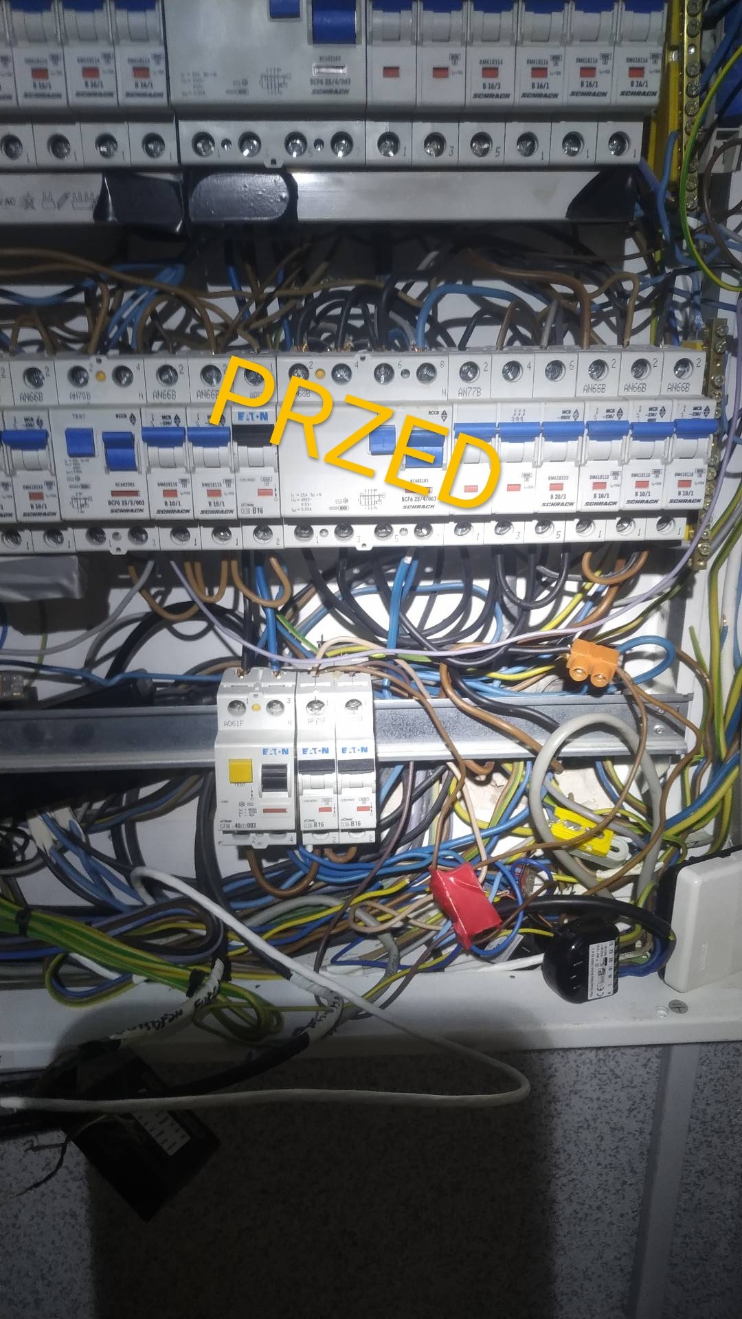 Otwarta skrzynka elektryczna z widocznymi bezpiecznikami marki Eaton i Schrack oraz splątanymi przewodami w różnych kolorach przed uporządkowaniem.