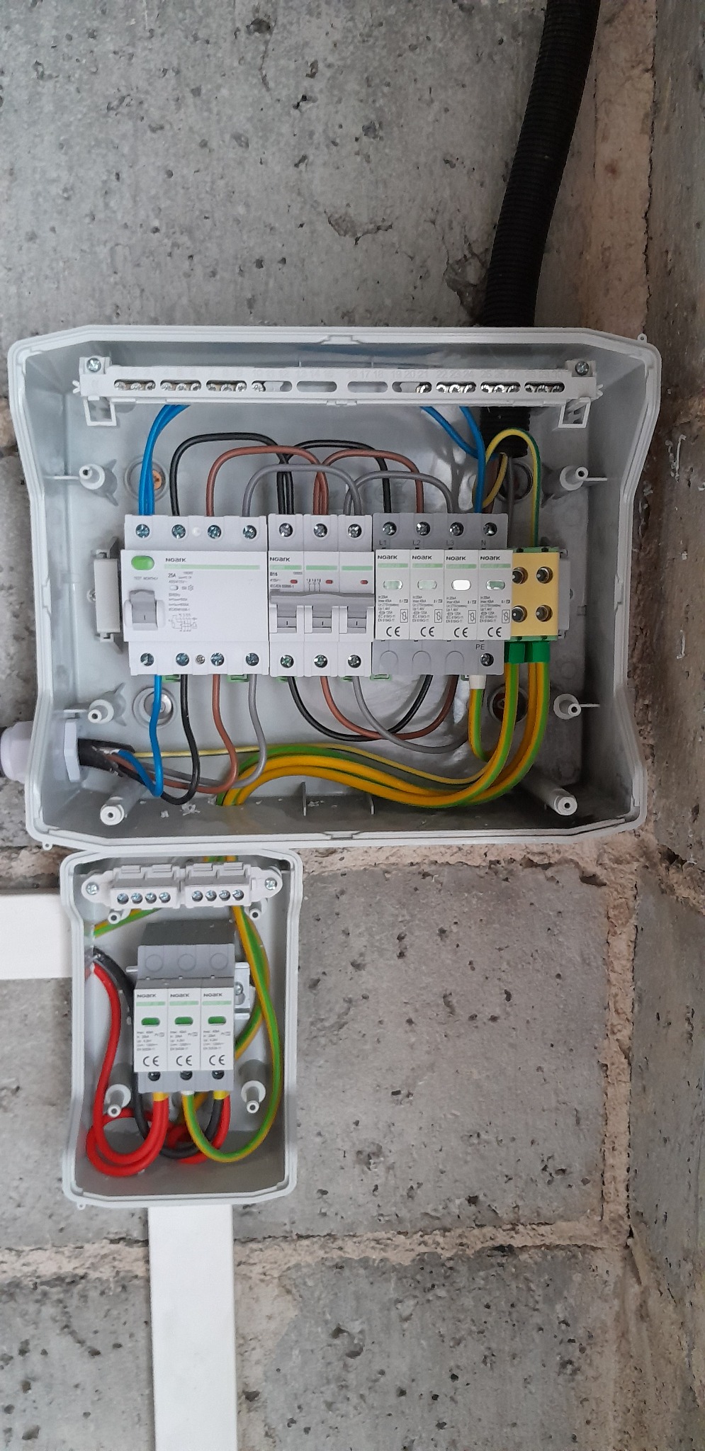 Rozdzielnica elektryczna z zainstalowanymi bezpiecznikami i przewodami w kolorach: niebieskim, czarnym, brązowym, żółto-zielonym, umieszczona na szarej, betonowej ścianie. Dodatkowa, mniejsza...