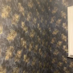 Ściana pokryta ciemną tapetą w złote liście palmy, widoczne gniazdko elektryczne i fragment zabezpieczonej podłogi.