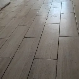 Ułożone podłużne płytki podłogowe imitujące drewno, widoczne krzyżyki dystansowe w spoinach, jasne światło słoneczne wpadające przez okno.