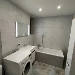 Nowoczesna łazienka z szarymi płytkami, podświetlanym lustrem, białą wanną obudowaną szarymi płytkami i pralką Whirlpool.