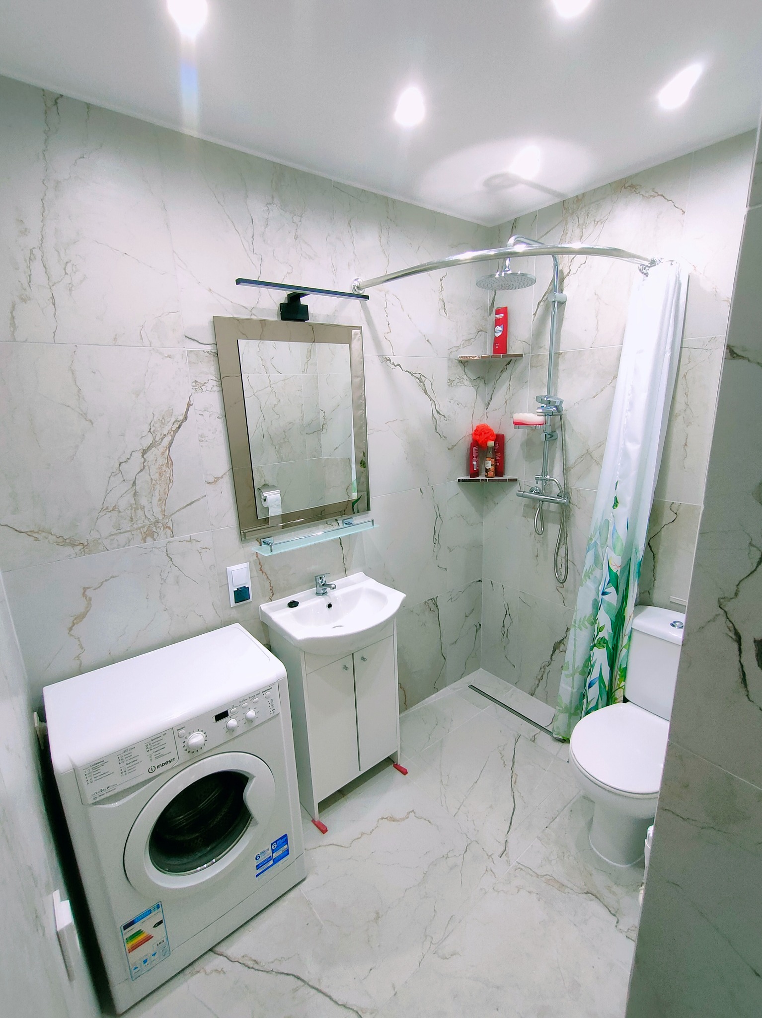 Jasna łazienka z marmurowymi płytkami, kabiną prysznicową, umywalką z szafką, toaletą i pralką Indesit.