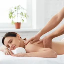 Klientka z zamkniętymi oczami relaksuje się podczas masażu pleców w jasnym gabinecie z rośliną w tle.