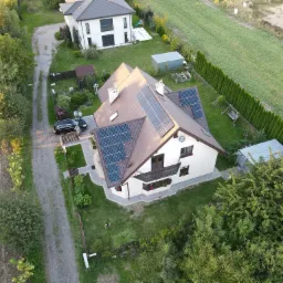 Widok z drona na dom jednorodzinny z brązowym dachem pokrytym panelami słonecznymi, na posesji zaparkowany czarny samochód, obok którego stoją dwie osoby.