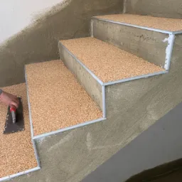 Prace wykończeniowe na schodach w Przecieszynie: aplikacja żywicznej posadzki z kruszywa kwarcowego za pomocą pacy. Widoczne metalowe profile krawędziowe.