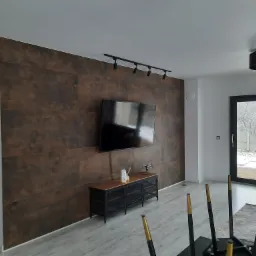 Nowoczesny salon z ścianą wykończoną panelami w odcieniach brązu, podwieszonym telewizorem i oświetleniem szynowym, widok na zasypany śniegiem ogród przez czarne drzwi balkonowe.