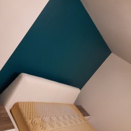 Emil Michalczak - Pomieszczenie na poddaszu z ukośnym sufitem, gdzie ściana pomalowana jest w dwie barwy: białą i turkusową, pod ścianą stoi rozłożony tapczan z materacem z pianki.