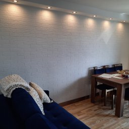 Emil Michalczak - Biała ściana z fakturą cegły podświetlona punktowymi lampami sufitowymi w pokoju z drewnianą podłogą, niebieską sofą i stołem z krzesłami.