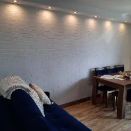 Biała ściana z fakturą cegły podświetlona punktowymi lampami sufitowymi w pokoju z drewnianą podłogą, niebieską sofą i stołem z krzesłami.