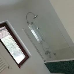 Nowoczesna łazienka z kabiną prysznicową z przezroczystego szkła, wykończona mozaiką w odcieniach zieleni, z oknem dachowym w drewnianej ramie i białymi ścianami.