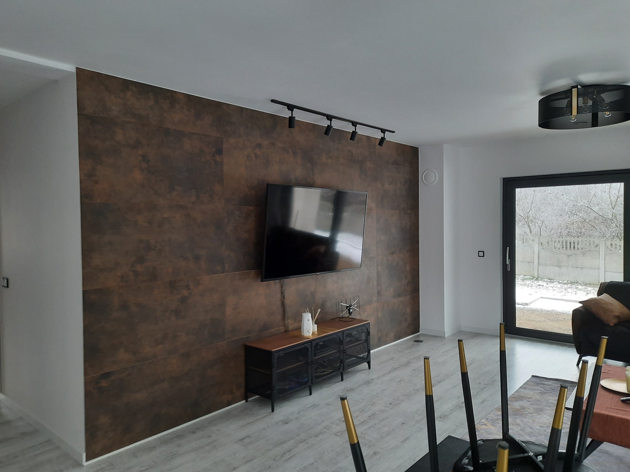 Nowoczesny salon z ścianą wykończoną panelami w odcieniach brązu, podwieszonym telewizorem i oświetleniem szynowym, widok na zasypany śniegiem ogród przez czarne drzwi balkonowe.