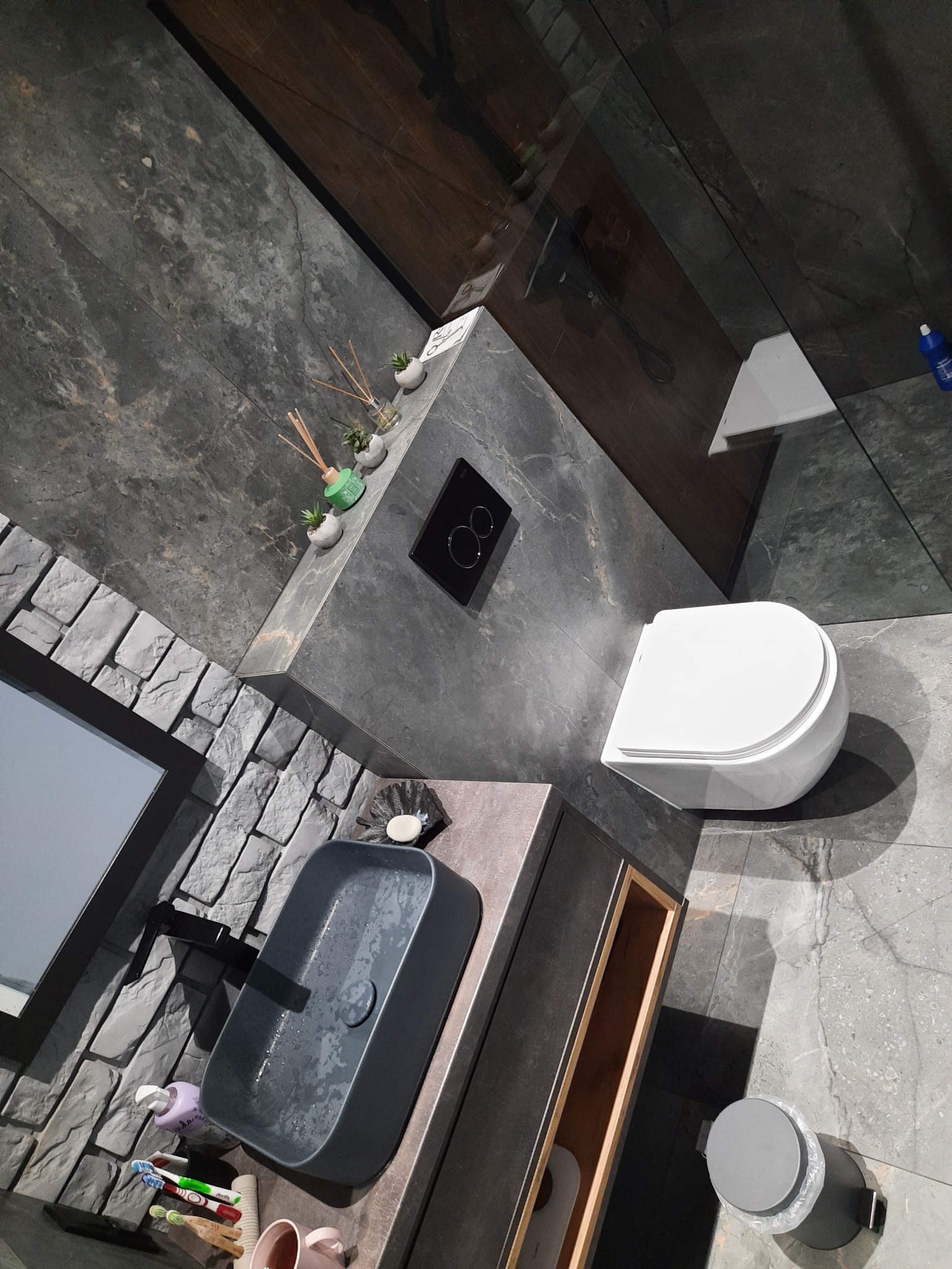 Nowoczesna łazienka z szarą mozaiką ścienną, umywalką nablatową i podwieszaną toaletą, uzupełniona o dekoracyjne akcenty zapachowe i minimalistyczne dodatki.