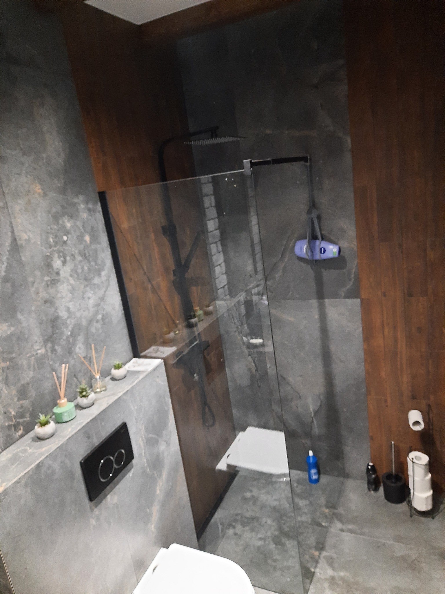 Nowoczesna łazienka z kabiną prysznicową typu walk-in, szare płytki imitujące marmur, drewniane panele na ścianie, wiszący sedes, dekoracyjne dyfuzory zapachowe i niebieski płyn do czyszczenia...